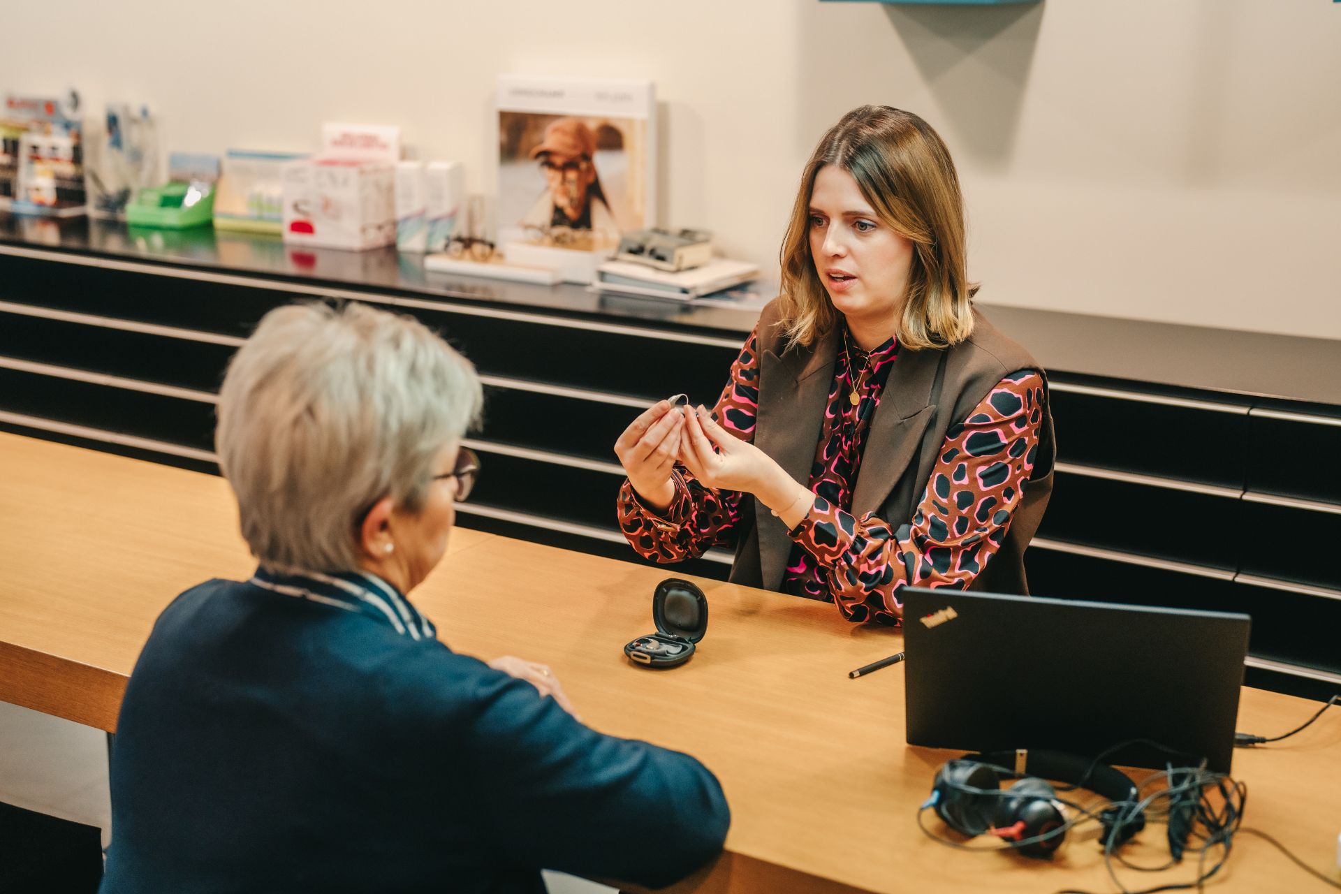 Charlotte heeft hoorapparaat vast en geeft uitleg aan klant aan tafel